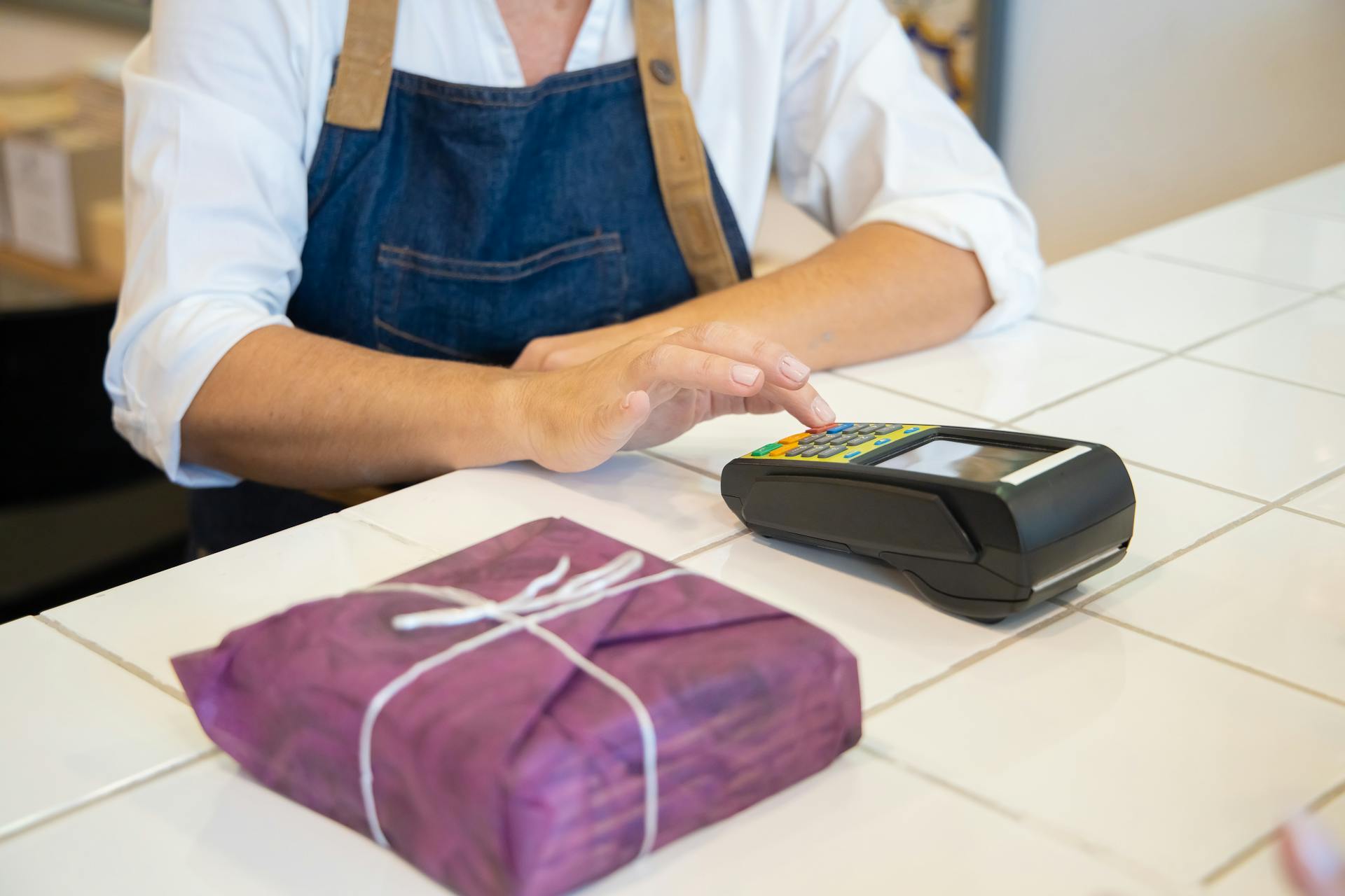 A person wearing a white shirt and blue apron stands behind a tiled counter, pressing buttons on a card payment terminal. A purple-wrapped package tied with white string is placed on the counter in front of them.